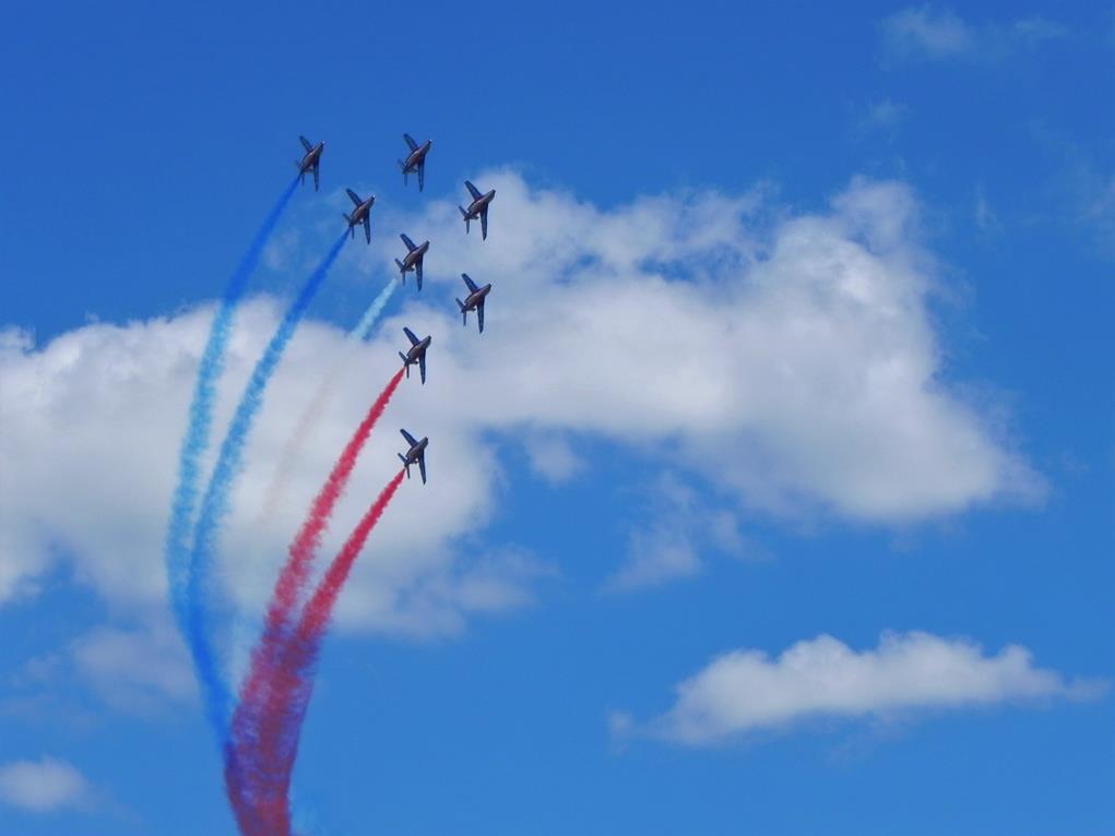 Un mouvement de la patrouille de France: sur un ciel bleu parsemé de nuages, les huit avions composant l'ensemble décrivent une pointe en arc de cercle, striant le ciel de trainées de couleurs à la verticale.