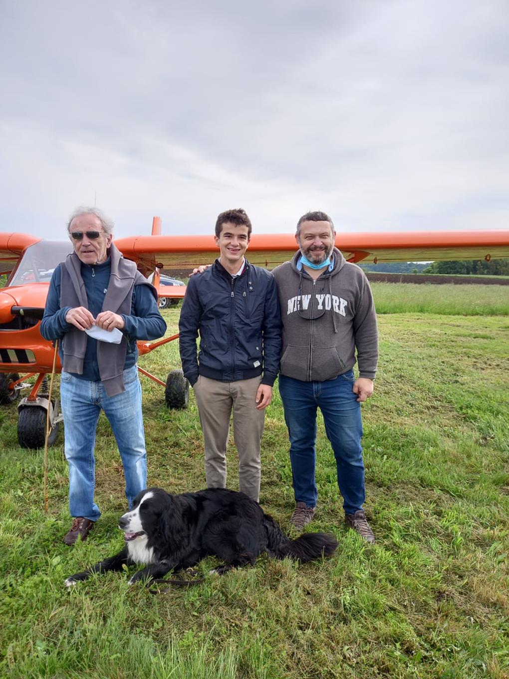 Devant l'avion en fond d'image, Martial et Jean-Claude posent avec Thomas ; grands sourires et accolades, le chien de Thomas à leurs pieds