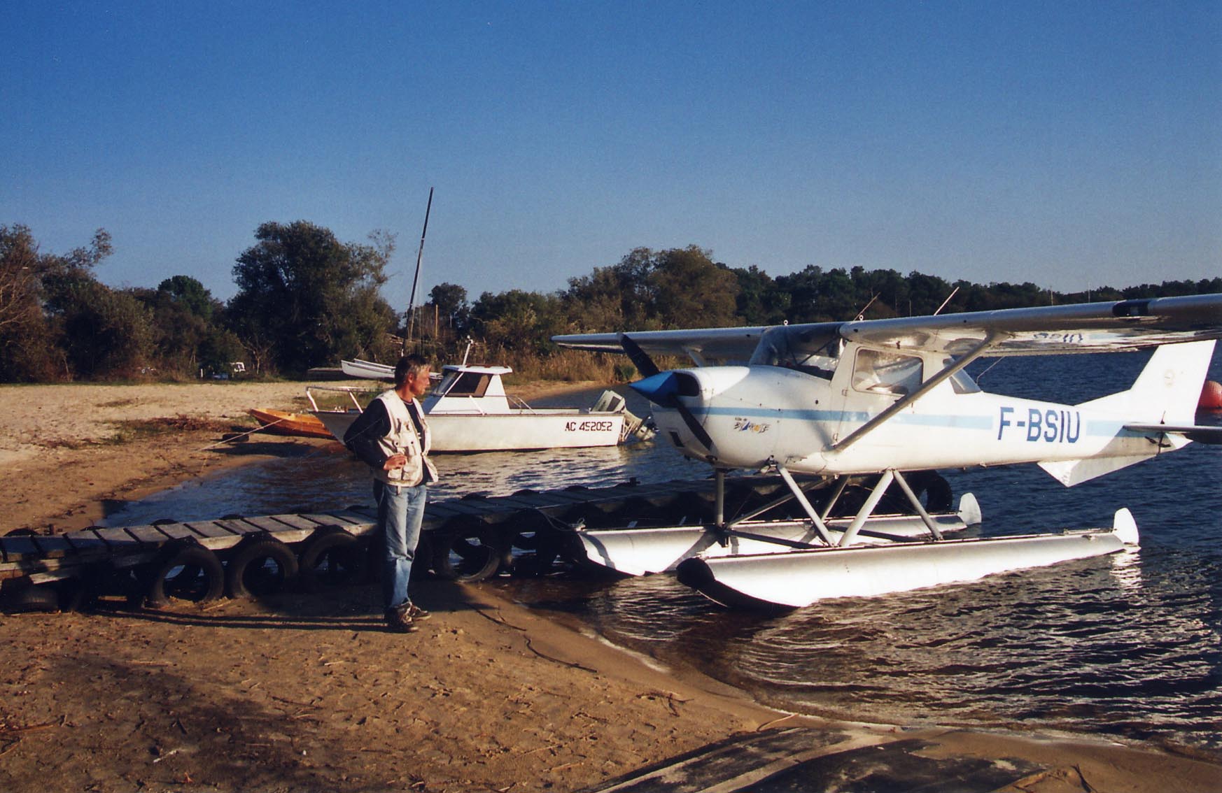 Dans un paysage de Lac - côtes sablées bordées d'arbres méditerranéens, pontons avançant dans l'eau et petits bateaux à quai - Eric contemple son F-BSIU en 2004
