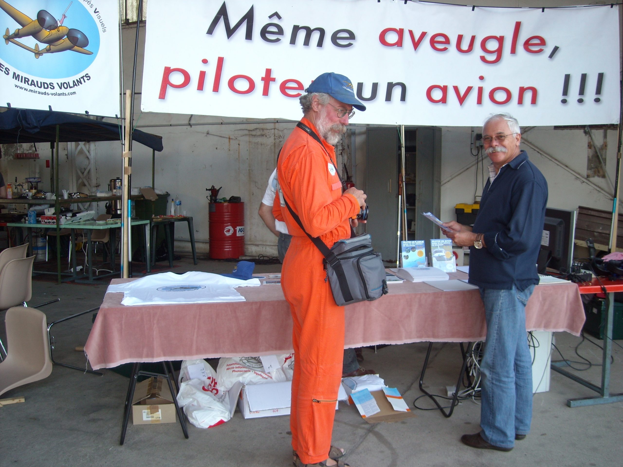 A Amiens, en 2009, Patrice, debout de profil dans sa combinaison orange, se tient devant le stand des Mirauds, au-dessus duquel une banderole flotte disant: "Même aveugle, pilotez un avion!"