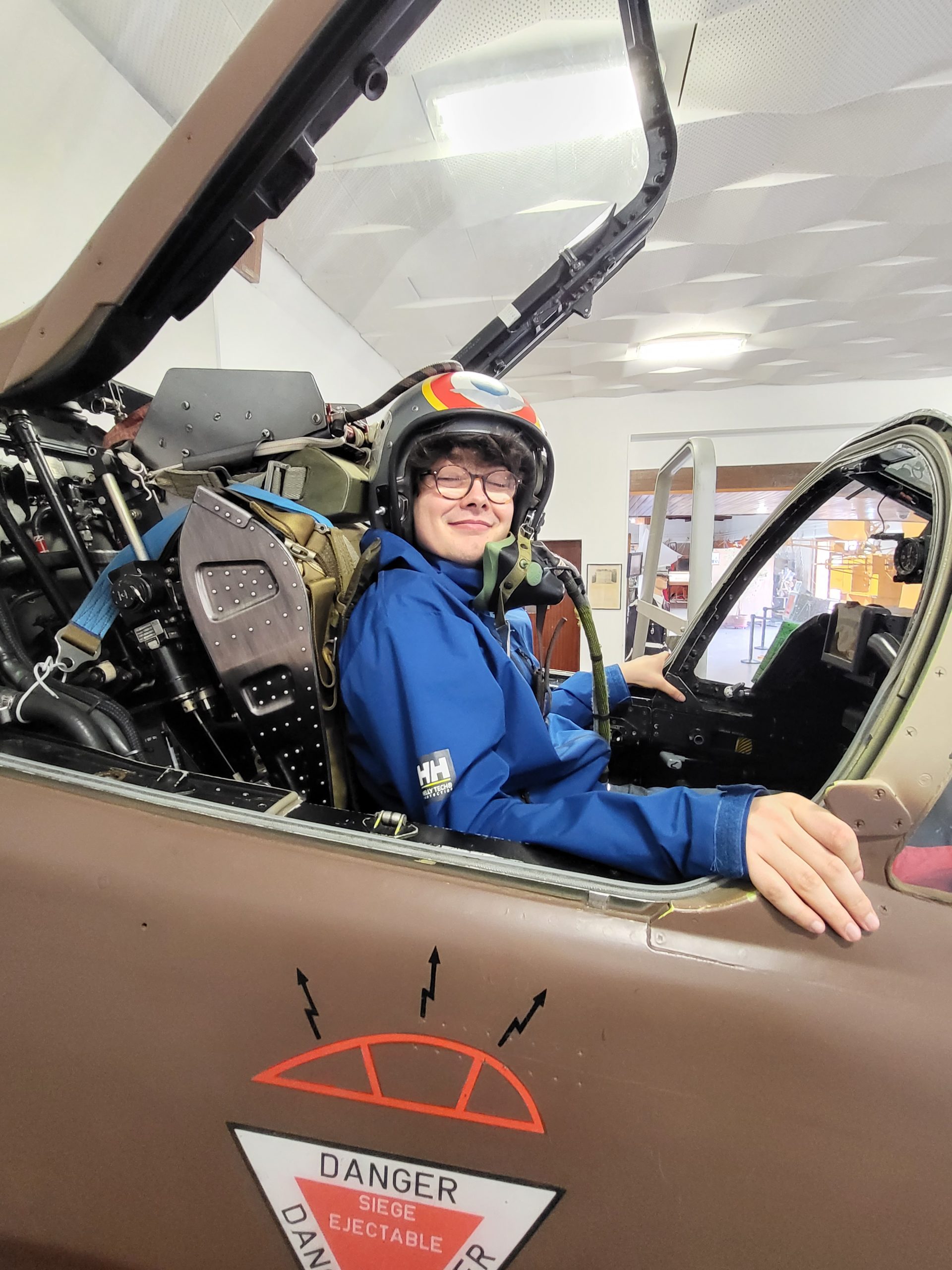 Portrait de notre adhérent Quentin dans le cockpit ouvert du Jaguar. Heureux, il porte tout l'équipement - casque, combinaison - comme en situation de vol. Sur le blanc de l'avion, un panneau indique "danger ! siège éjectable".