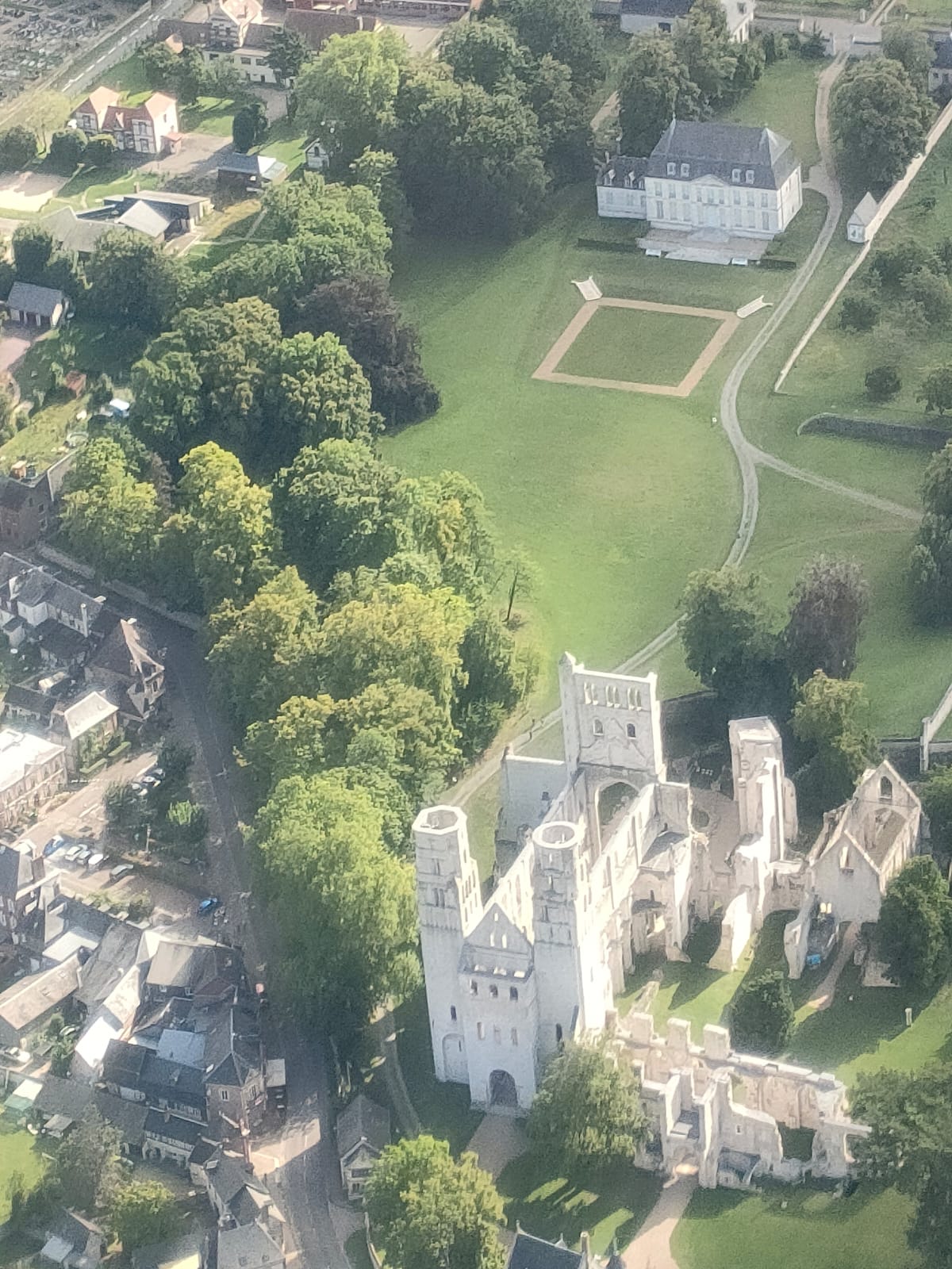 L'abbaye de Jumièges vue du ciel, comme posée au milieu d'une maquette. Les tours d'architecture romane, s'élèvent au milieu des arbres, tandis que le reste du paysage est ville d'un côté, étendues gazonnées de l'autre.