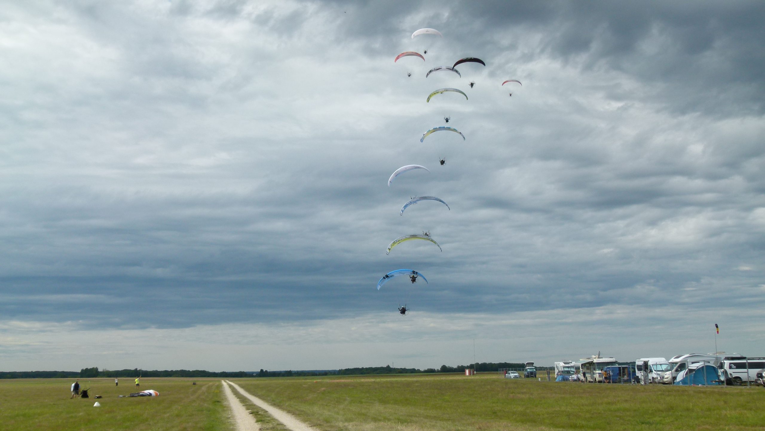 Démonstration de parachutisme. Sur un ciel cotonneux, une douzaine de parachutes descendant en colonne vers le sol herbeux amorcent leur atterrissage .