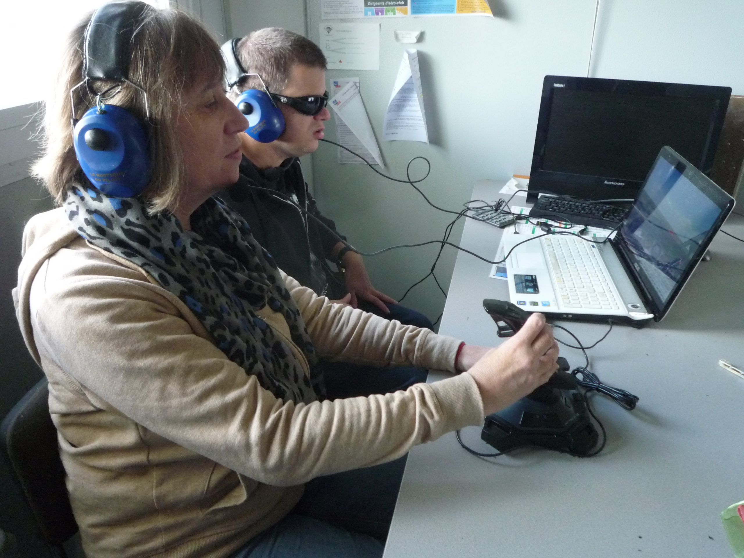 Dans une salle, Christine et Kevin utilisent le simulateur de vol. Chacun porte un casque audio relié aux écrans ; Christine manipule le joystick pour les commandes.