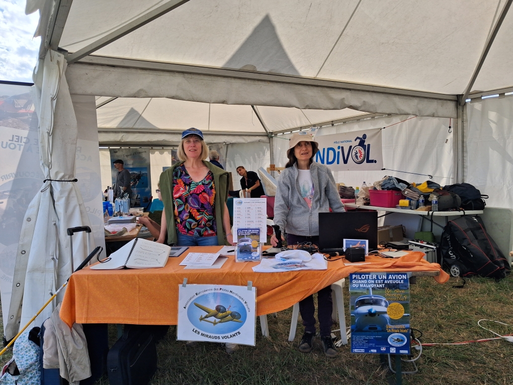 Vue de face sur le stand de l'association. Isabelle et Christine, souriantes, debout derrière la table et sous le chapiteau. Derrière, d'autres stands. Sur la table, des informations et logo de l'association ainsi que le simulateur de vol.