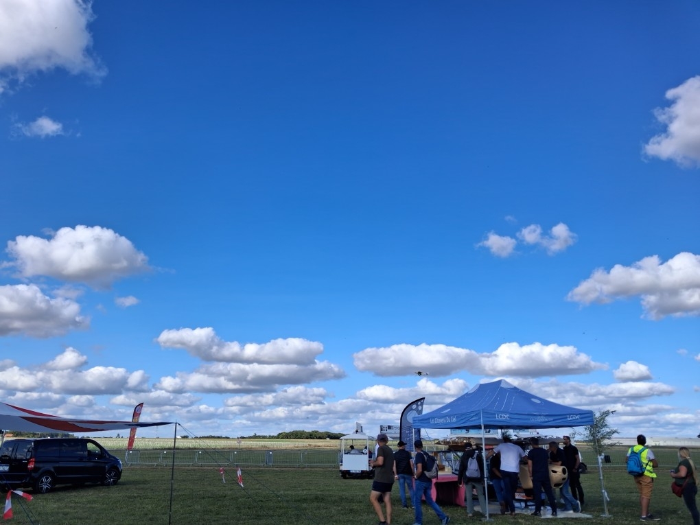Vue globale sur le salon. En extérieur, sur fond d'un grand ciel bleu parsemé de quelques nuages seulement. le terrain est plat est engazonné. A gauche et à droite de l'image des stands sous des barnums avec des visiteurs.