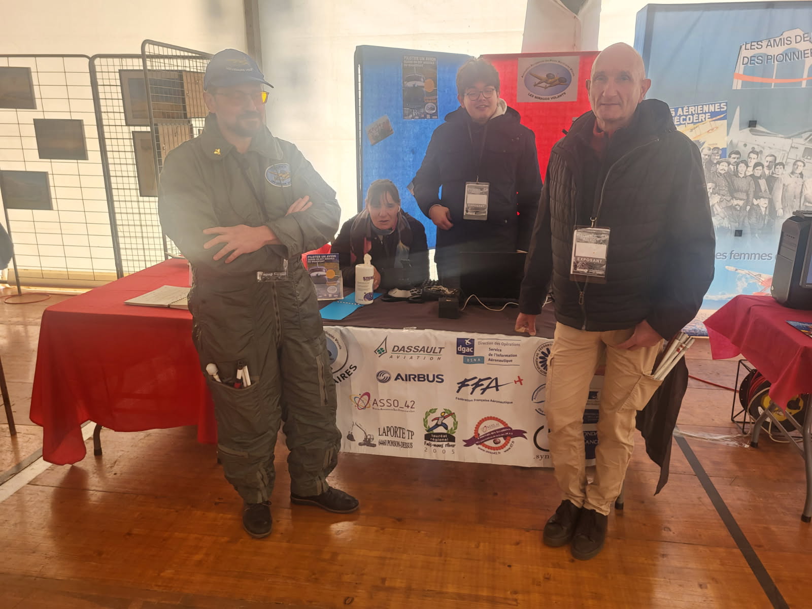 Quatre personnes devant et derrière un stand d’exposition. À gauche, un homme porte une combinaison de vol verte et une casquette bleue, bras croisés, avec des outils dans sa poche. Deux personnes sont derrière la table : une femme accroupie, tenant une bouteille, et un homme souriant avec un badge autour du cou. À droite, un homme chauve porte une veste noire et un pantalon beige, une main dans la poche, un badge autour du cou. La table est recouverte d’une nappe blanche affichant des logos de sociétés aéronautiques comme Airbus, Dassault et FFA. Derrière, un panneau bleu et rouge ainsi que des affiches sur le thème de l’aviation. Le sol est en bois, l’ambiance semble être celle d’un salon ou forum.