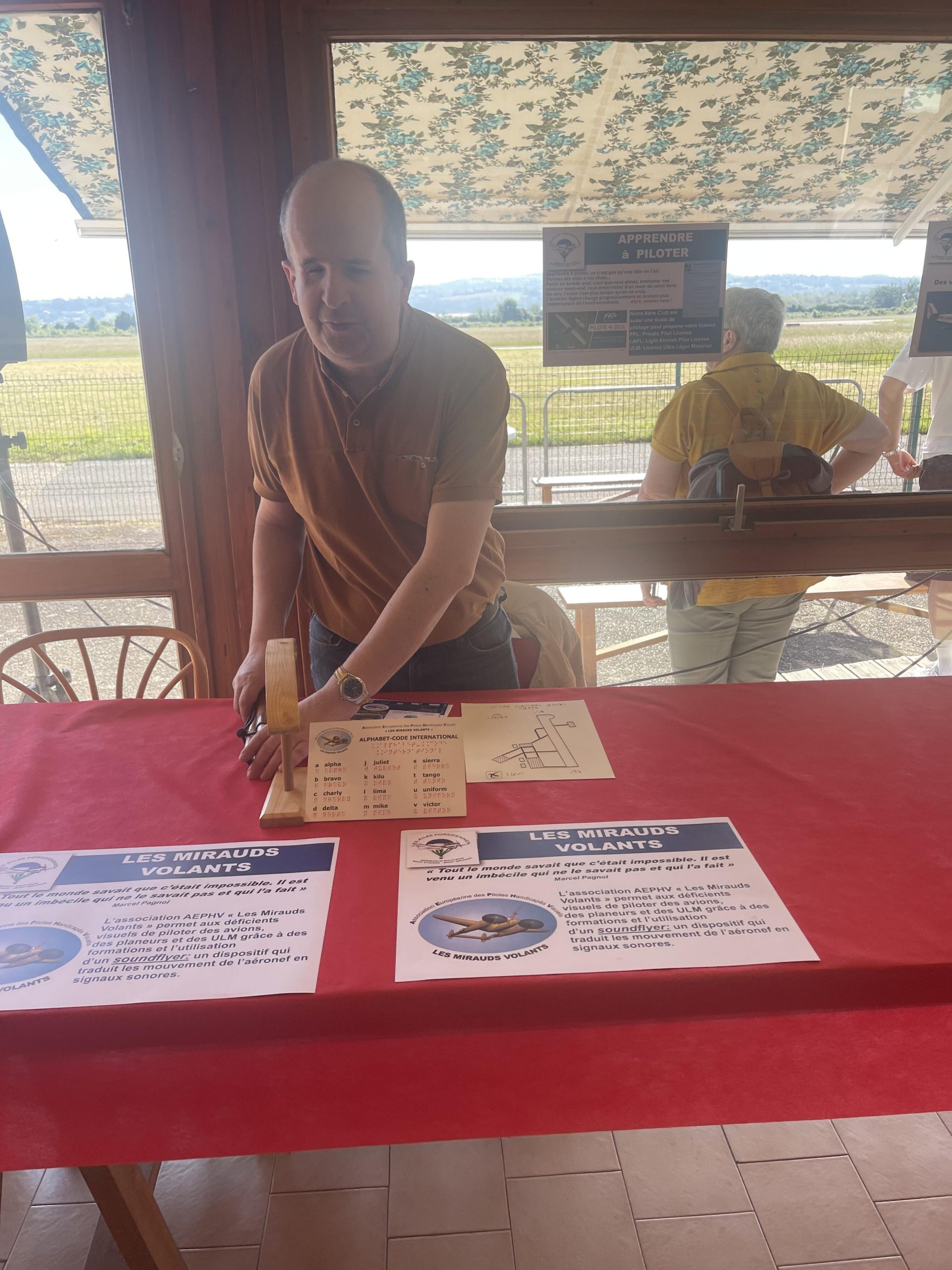 Thomas, tout sourire, met en place les éléments de présentation de son stand pour les journées portes ouvertes des Ailes Foréziennes. En fond, l'extérieur de l'aéroclub sous le soleil.