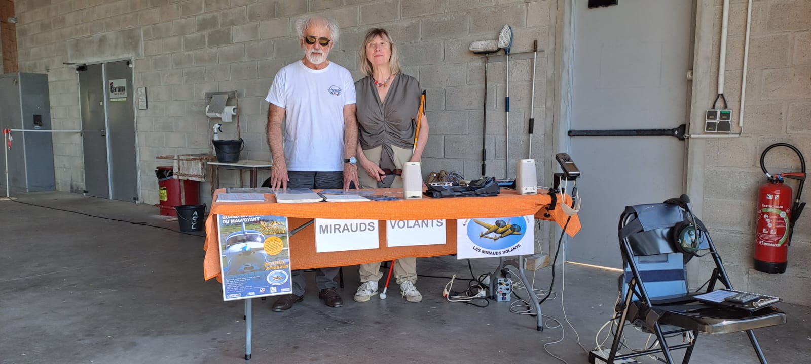 Deux de nos membres posent au stand des Mirauds volants pour ces journées portes ouvertes. Ils sont debout à une table avec affiches et dépliants. Devant la table, une chaise est installée avec tout les éléments du soundflyer pour une démonstration.