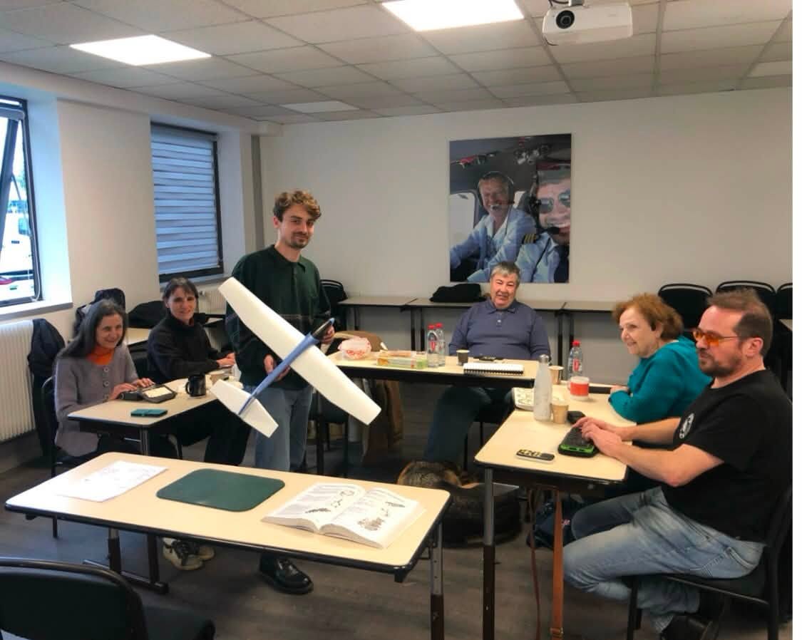 Dans une salle de classe, nos 5 apprentis sont assis à des tables disposées en U. Au centre, leur formateur avec une maquette d'avion.